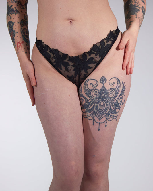 Person wearing black lace lingerie with visible tattoos on a plain background